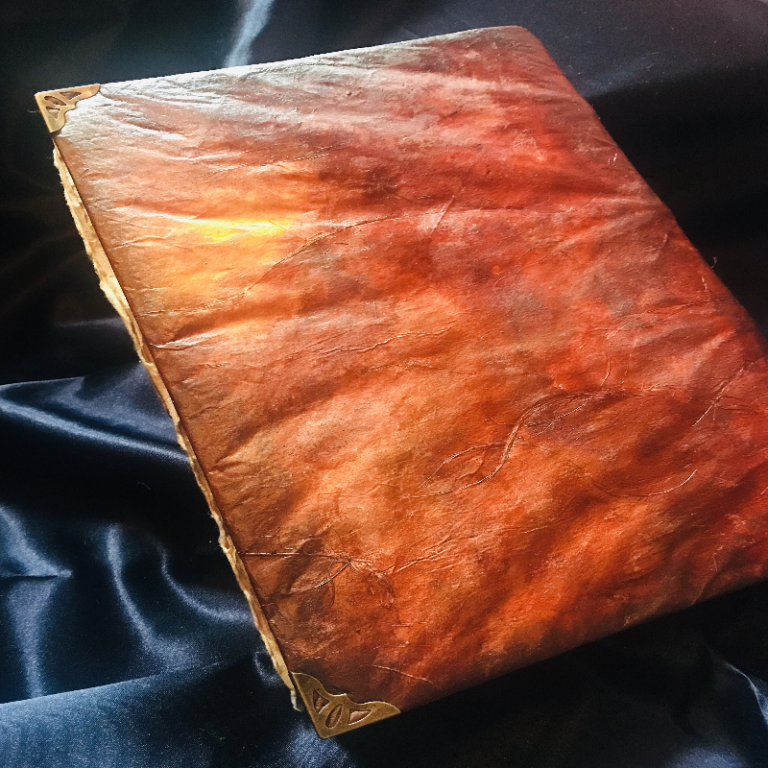 A closed, textured like- leather-bound book with ornate corner details rests on a dark, silky fabric surface.